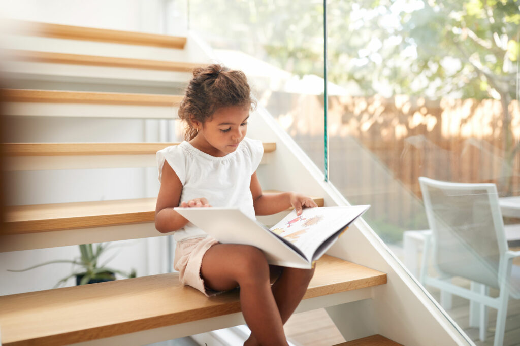 young african american girl reads a story book on the stairs in her home