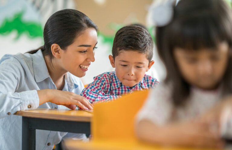 A reading specialist teacher engages a student one-on-one, as he reads a book on the desk