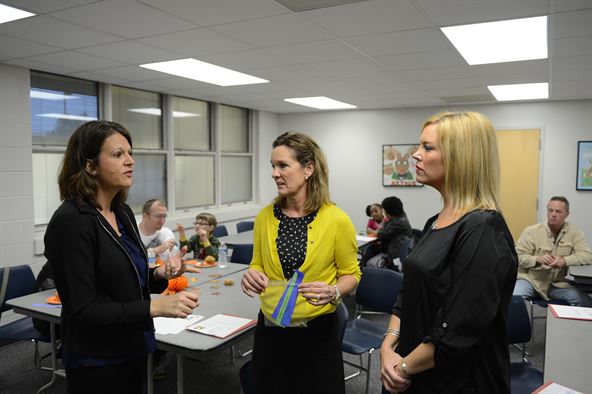 Photo of three women talking while kids are sitting at desks behind them.