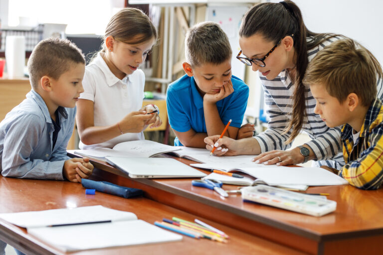 Surrounded by workbooks and pencils, a group of students focus on a teacher's explanation of phonics