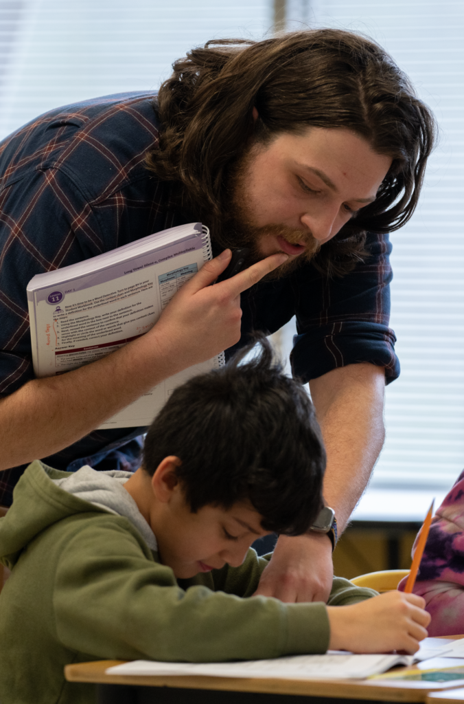 A male teacher holds a teachers' edition while helping a student who is writing in his reading workbook