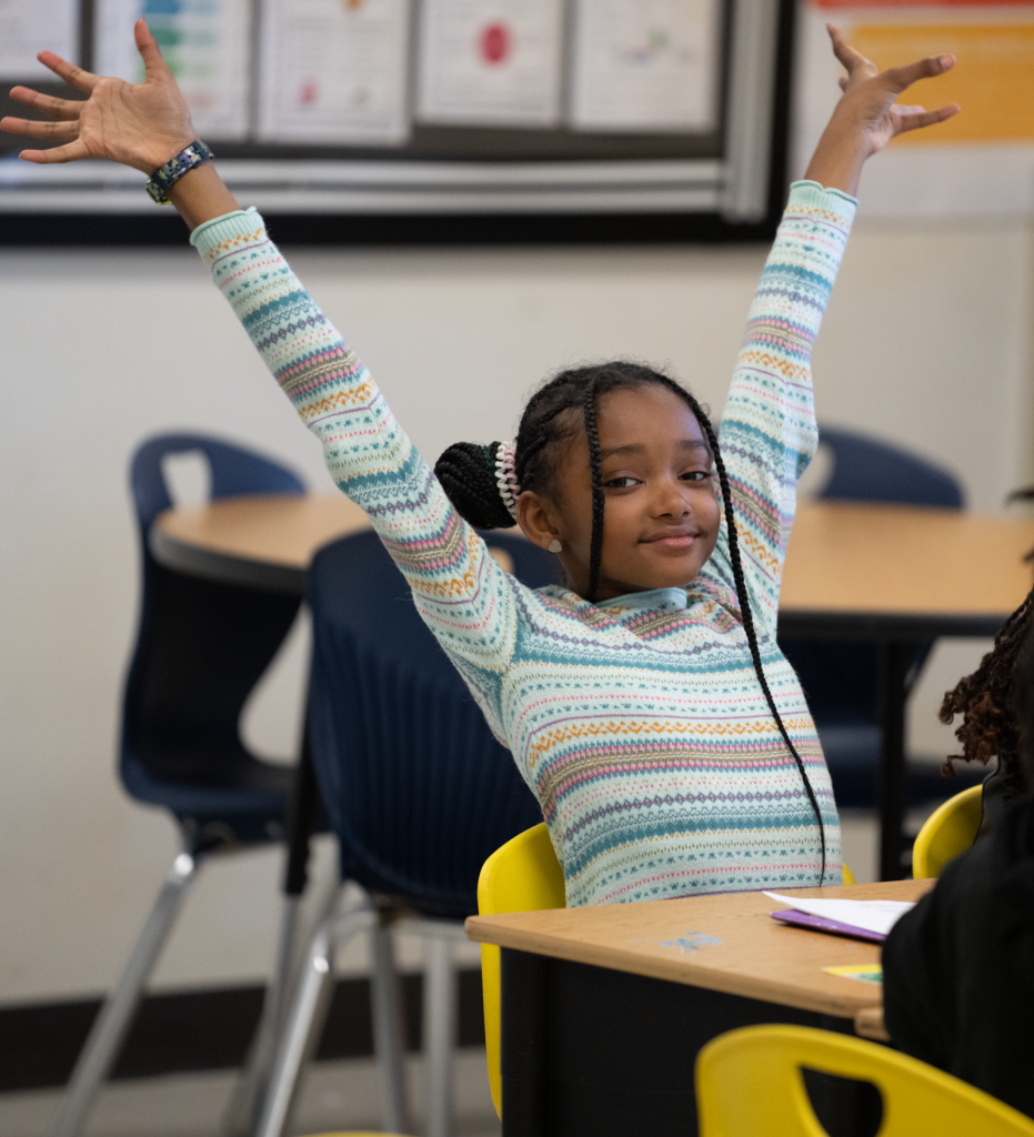 A happy elementary school girl stretches her arms up in her classroom