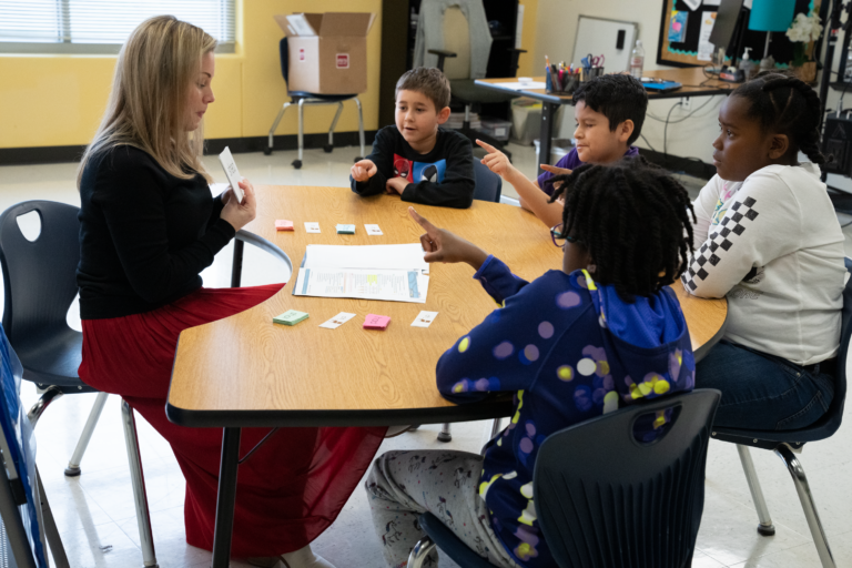 Students sitting at a semi-round deck with a teacher in the middle reviewing routine cards.