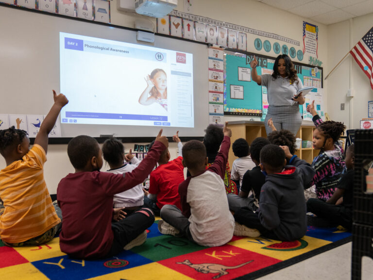 Reading students raise hands as their literacy teacher explains items show on a smart board