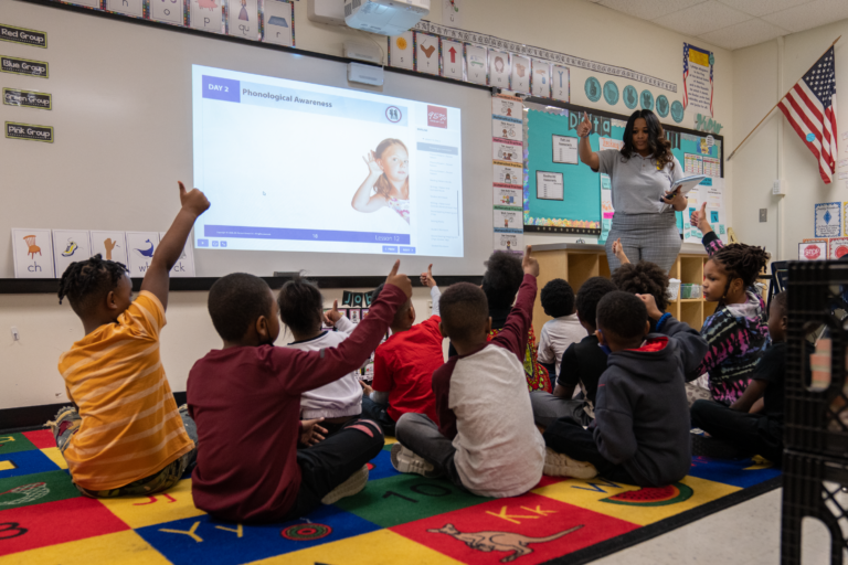 Children sitting on a classroom rug giving thumbs up to their teacher.