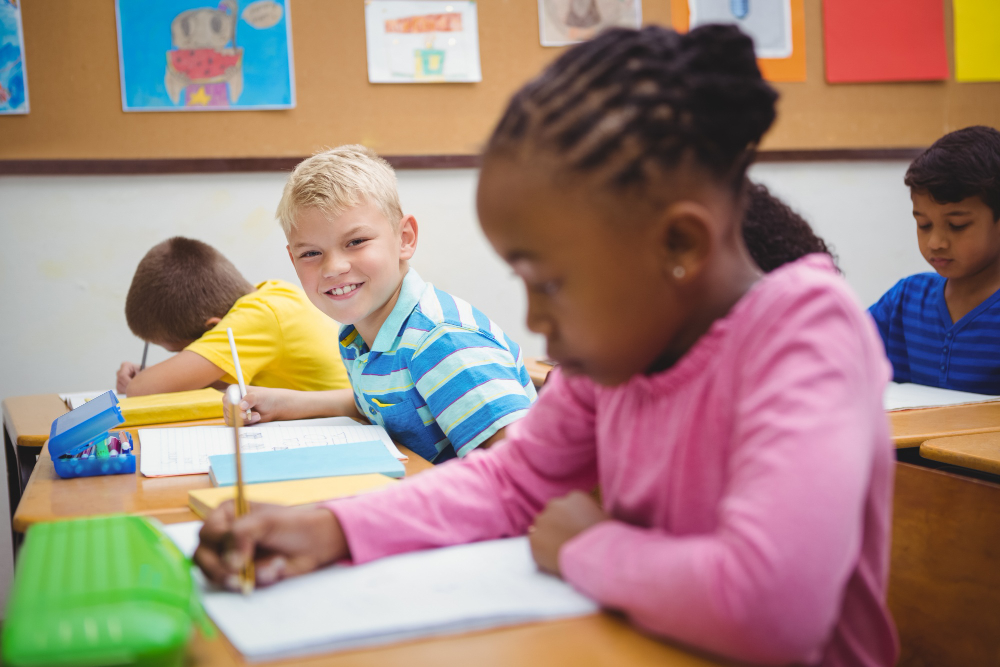 An elementary school girl is hard at work in her phonics workbook while a fellow student smiles