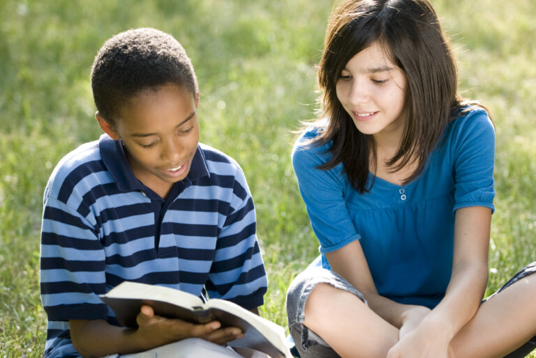middle school boy reading a book to a girl student outside