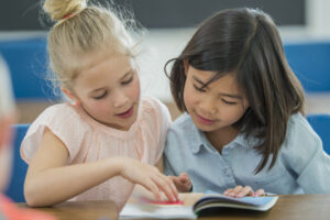 Two elementary school girls work together to sound out words in their book at school.