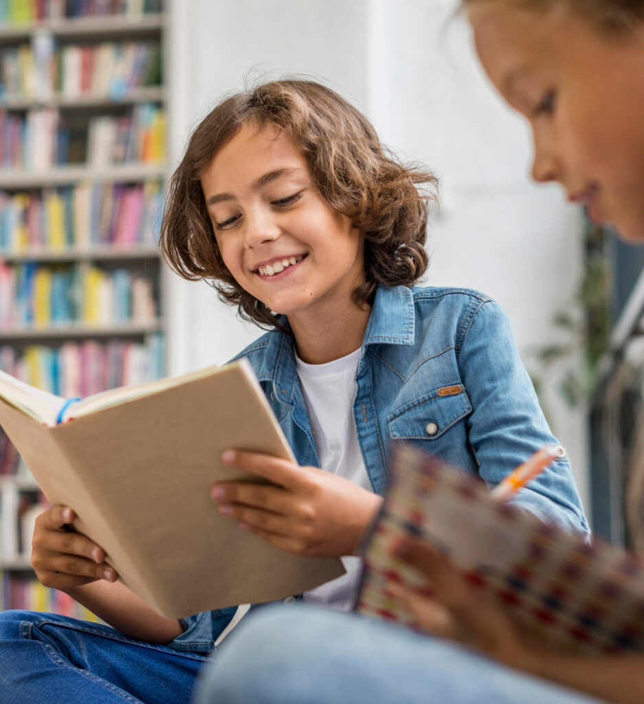 A middle school boy smiles while reading a book in a library, while a middle school girl writes in a journal