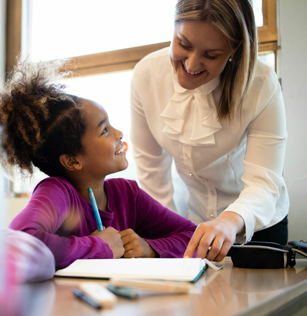 A teacher gives one-on-one attention to a student using a pencil and workbook