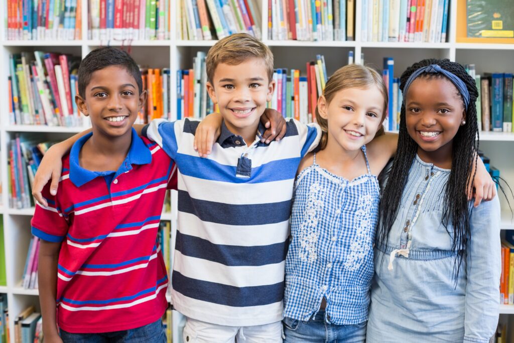 Four older elementary school students smile and embrace in front of books in the library