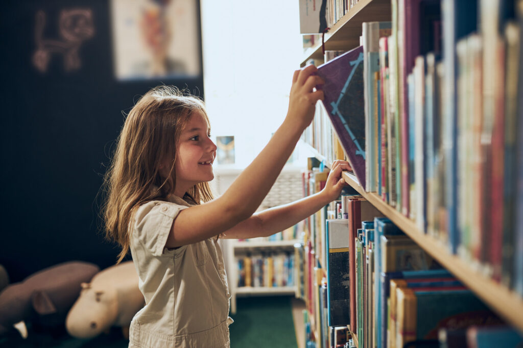 elementary school girl smiles and pulls book from bookshelf in school library