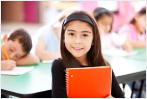 Latina elementary school student holds notebook while smiling in grade school classroom.