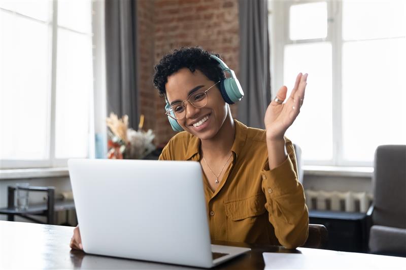 African-American female educator waves happily to laptop camera while accessing online webinar training from her home office.