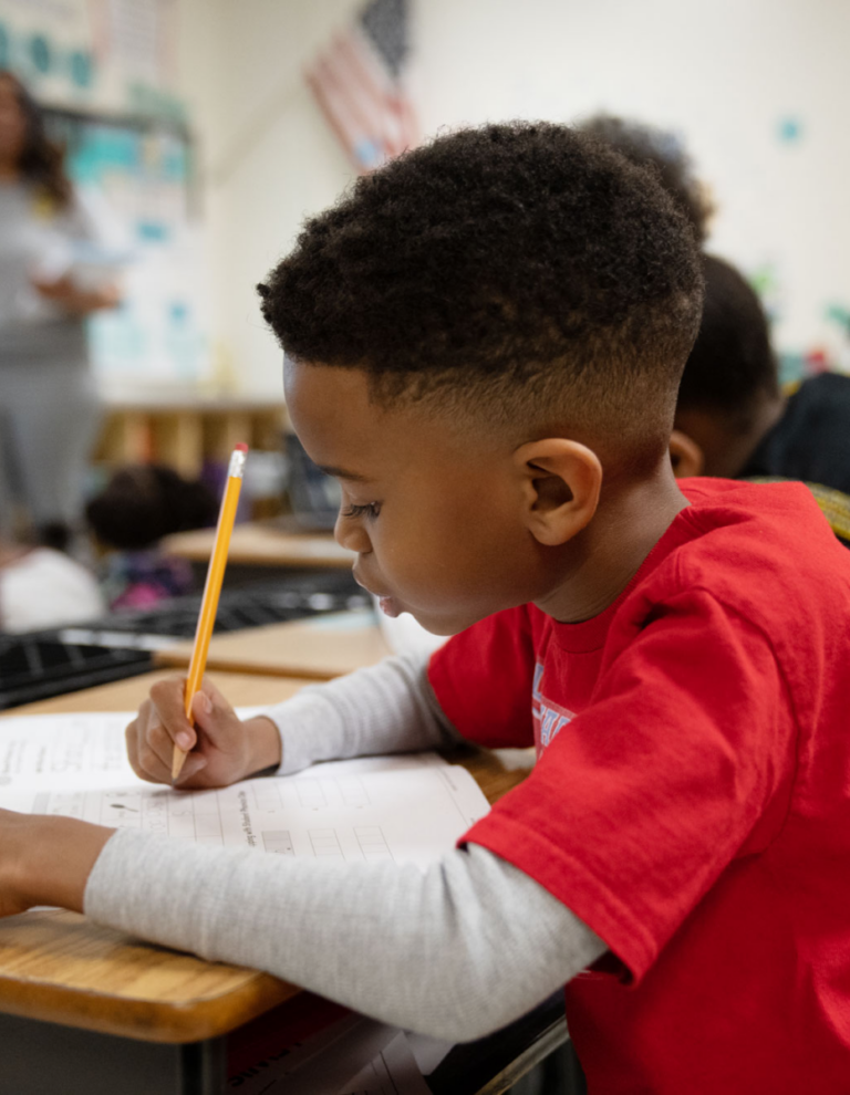 Student writing in classroom with teacher in background