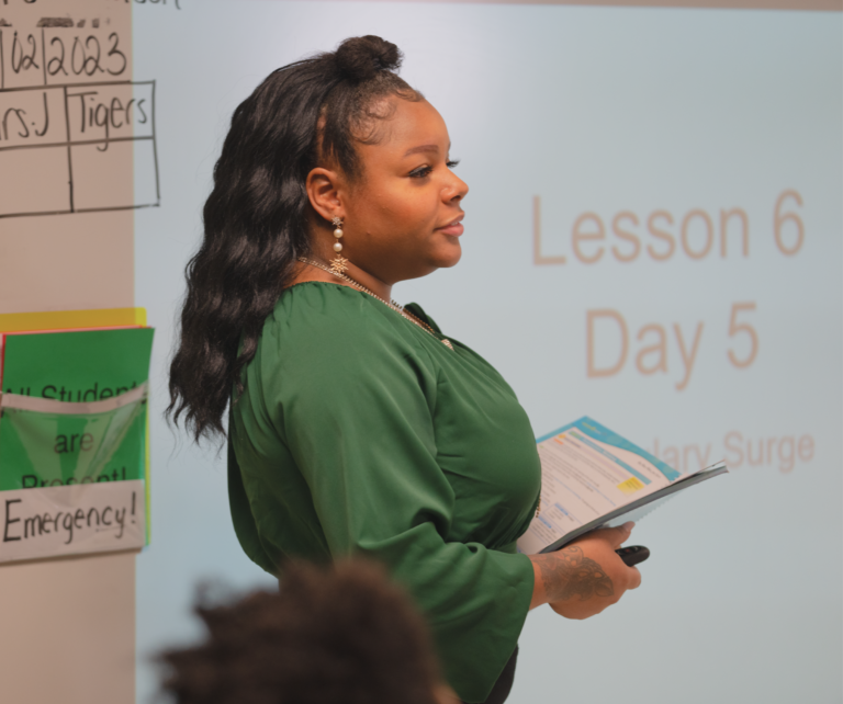 Teacher in green with book presenting lesson in classroom