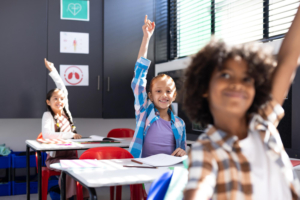 Diverse happy children sitting at desks raising hands in elementary school
