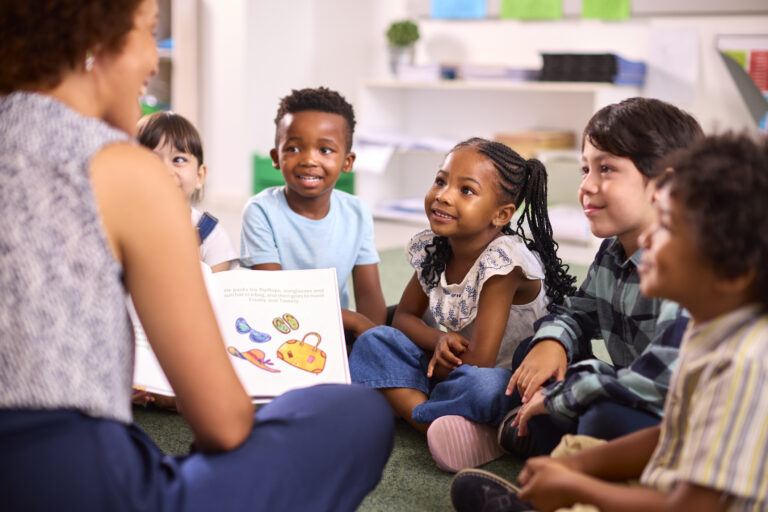 Female Teacher Reads To Multi-Cultural Elementary School Pupils Sitting On Floor In Class At School