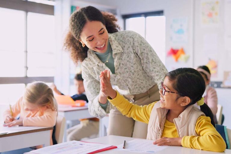 Teacher woman, high five girl and classroom with achievement, success and mentorship for learning. Education, development and students with goals, knowledge and books with celebration at school desk