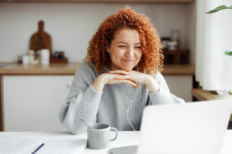 Female freelancer having video conference with her employer discussing new project details, listening to him attentively in headphones smiling at web camera, putting folded hands under chin