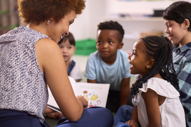 Female Teacher Reads To Multi-Cultural Elementary School Pupils Sitting On Floor In Class At School
